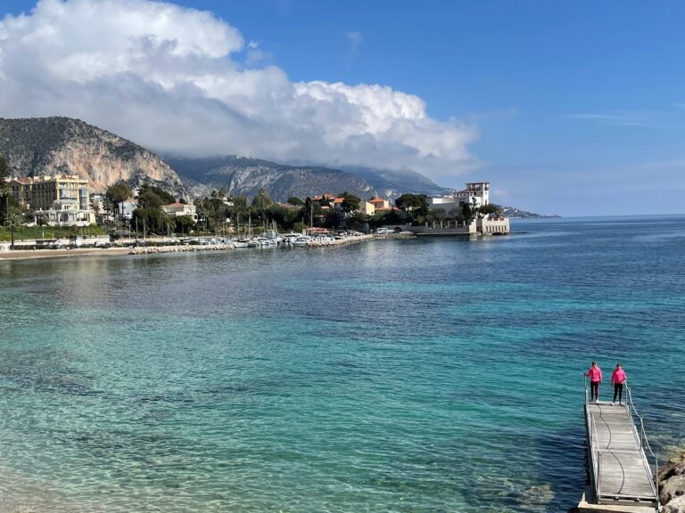 A scenic coastal view captures two people standing on a dock, overlooking the clear turquoise waters of the French Riviera. The shoreline boasts charming buildings and trees, set against green hills and a partly cloudy blue sky. Visiting the French Riviera in the off season may be one of the best times to go!