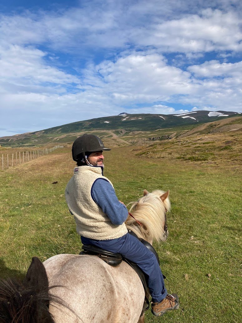 A person wearing a helmet and fleece vest rides a light-colored horse in a grassy, open landscape with rolling hills and patches of snow under a partly cloudy sky.