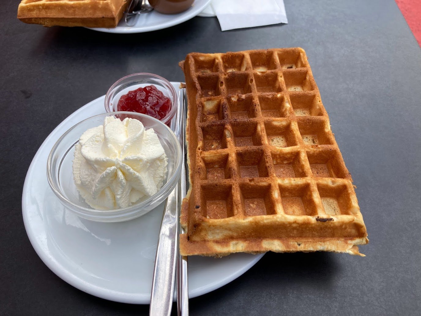 A plate with a rectangular waffle, a small bowl of whipped cream, and a small bowl of red jam on a dark table. A fork and knife are placed on the plate, and another plate is partly visible in the background.