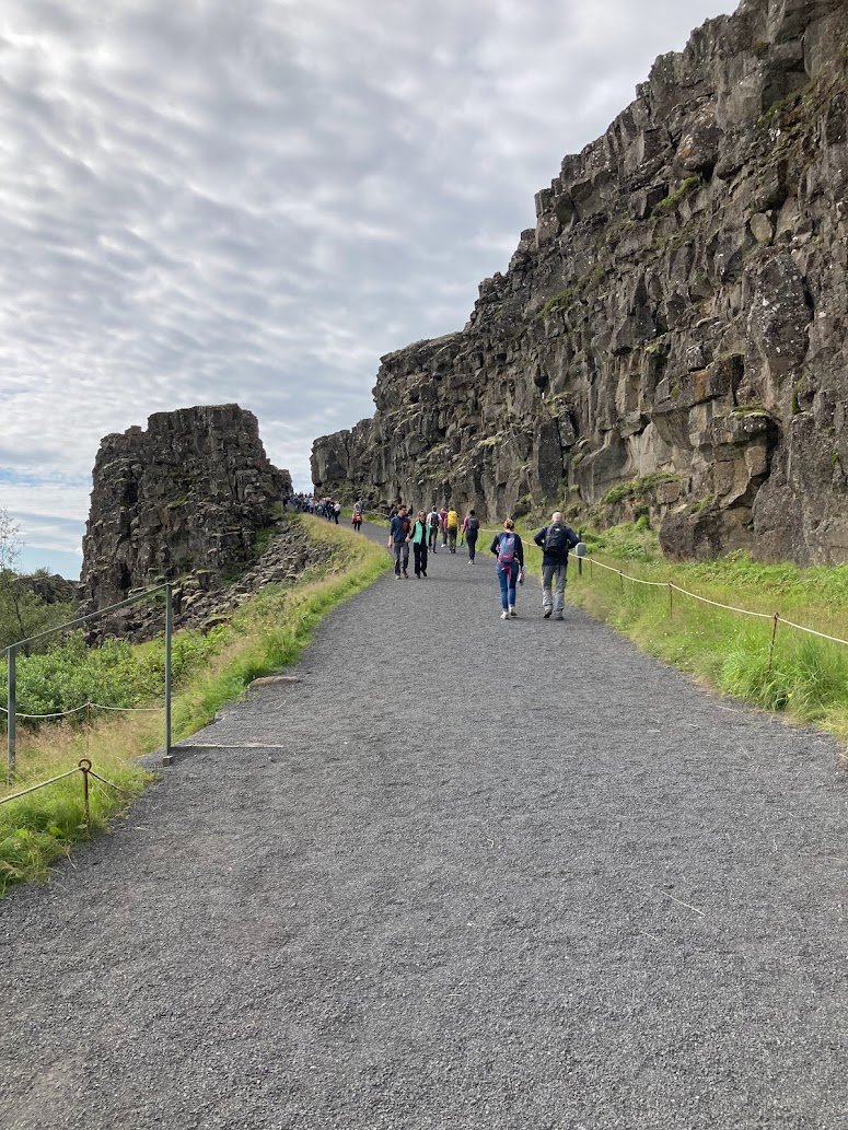 People walk along a gravel path bordered by green grass and rocky cliffs under a cloudy sky. The path is lined with a simple rope fence, and the scenery appears rugged and natural.