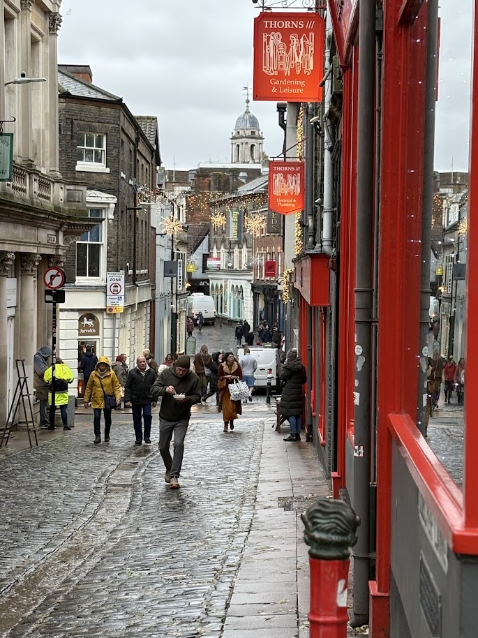 A narrow cobblestone street in a town with people walking. Shops with red facades line the street. In the distance, a domed building is visible. The street is decorated with lights against a cloudy sky.