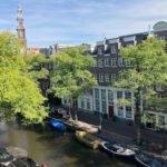 Some Amsterdam travel tips: Walk along the canals in Amsterdam lined with boats, trees, and traditional Dutch brick buildings under a blue sky—one of the charming sights you'll see when visiting Amsterdam. A tall church tower rises in the background.