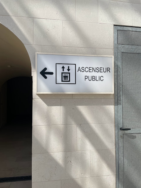 Sign on a stone wall with an arrow pointing left, symbol of an elevator, and text ASCENSEUR PUBLIC, pointing to the public elevator—a handy tip when you travel the French Riviera in the off-season. Shadow patterns dance on the wall and floor.