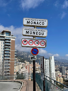 A road sign marks the entrance to Monaco, showcasing both French and a regional language. It displays traffic restrictions and a 50 km/h speed limit. In the background, tall buildings and a mountainous landscape invite you to travel the French Riviera in the off season.