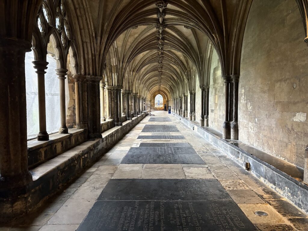 A historic stone cloister with vaulted ceilings and arched windows on the left stands as a must-see for those wondering where to go in Norfolk. Sunlight filters in, illuminating the stone floor with engraved slabs. The passageway extends into the distance, creating a sense of depth and history.