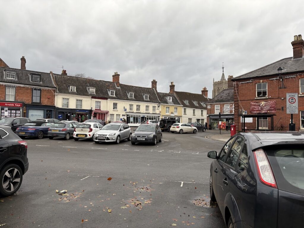 A cloudy day at a town square in Aylsham, Norfolk with parked cars and red brick buildings. The scene, a hint of where to go in Norfolk, includes businesses and homes, with a church spire visible in the background. Fallen leaves are scattered on the ground.