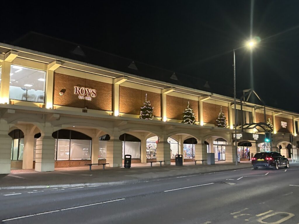 Night view of a lit-up brick building with arched entrances labeled ROYS, one of the charming spots on the list of where to go in Norfolk. Christmas trees with lights are visible in the windows. A street with road markings and a passing car is in the foreground.