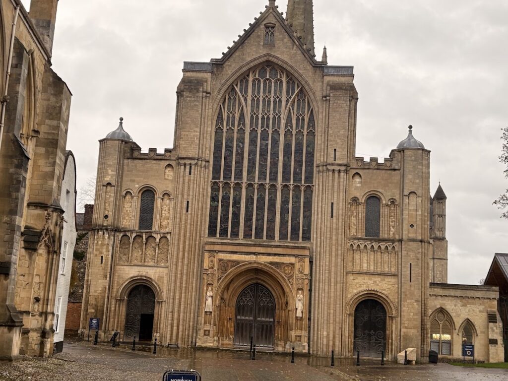 A historic stone cathedral in Norfolk, England, stands solemnly on a cloudy day, showcasing large arched windows and ornate architectural details. The facades two towers and pointed roof loom over the wet ground, hinting at recent rain—a quiet charm during off-season travel.