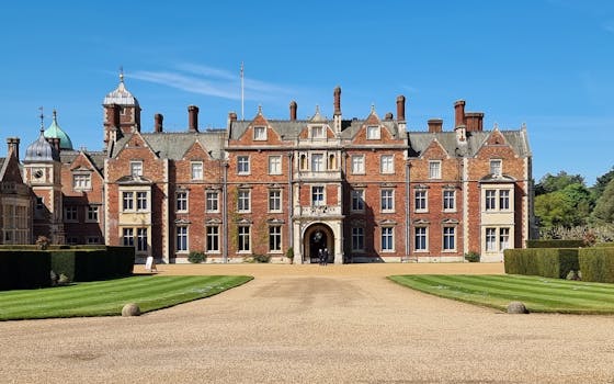 Sandringham House is a large, historic brick mansion in Norfolk, England, featuring ornate details, multiple chimneys, and a central archway. The building is surrounded by well-manicured lawns and hedges under a clear blue sky.