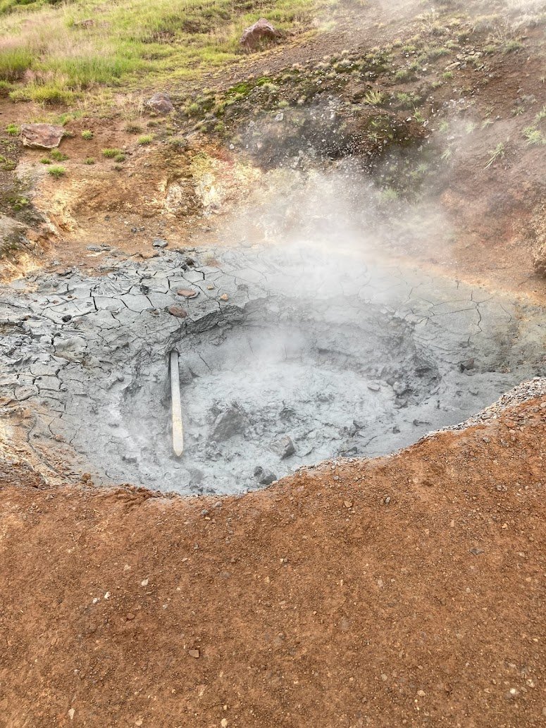 A steaming gray mud pool with cracked edges in a geothermal area, just off the Ring Road, surrounded by reddish-brown soil and sparse green grass. A wooden stick lies partially submerged in the mud—a unique sight while camping in Iceland.