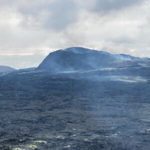 A misty landscape with a dark, rocky terrain in the foreground, faint blue smoke rising, and a distant mountain under a cloudy sky evokes the wild beauty of camping in Iceland along the iconic Ring Road.