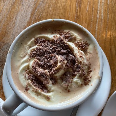 A cup of hot chocolate topped with whipped cream and chocolate shavings, served on a white saucer on a wooden table—perfect after a day of camping in Iceland or exploring the Ring Road.