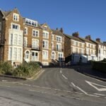 A row of tall, brown brick Victorian-style houses with white window frames lines a curving street under a clear blue sky—classic scenery for those seeking unique things to do in Norfolk. A red postbox and some parked cars are visible near the houses.