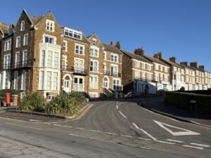 A row of tall, brown brick Victorian-style houses with white window frames lines a curving street under a clear blue sky—classic scenery for those seeking unique things to do in Norfolk. A red postbox and some parked cars are visible near the houses.