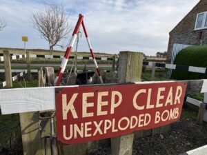 A red and white sign reading KEEP CLEAR UNEXPLODED BOMB is attached to a wooden fence, with warning barriers and a tripod marking a cordoned-off area in a rural setting.