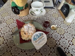 A plate with a scone dusted with powdered sugar, a knife, a small jar of jam, and a packet of clotted cream on a lace-patterned tablecloth. A teacup, saucer, and utensils are in the background.