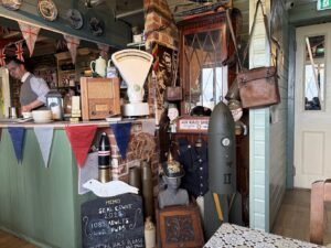 A vintage-themed café interior with bunting, old-fashioned scales, posters, a World War II bomb replica, helmets, a chalkboard sign, and various retro items scattered around the counter and walls.