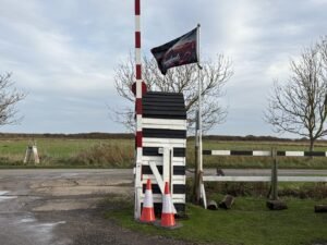 A black and white striped wooden gate structure stands by a rural road with three orange traffic cones in front, a worn flag waving above, and bare trees and green fields in the background.