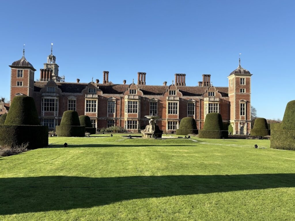 A large historic brick mansion with tall chimneys and towers, perfect for day trips from Norwich England, stands behind neatly trimmed hedges and a wide, sunlit lawn under a clear blue sky.