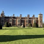 A large historic brick mansion with tall chimneys and towers, perfect for day trips from Norwich England, stands behind neatly trimmed hedges and a wide, sunlit lawn under a clear blue sky.