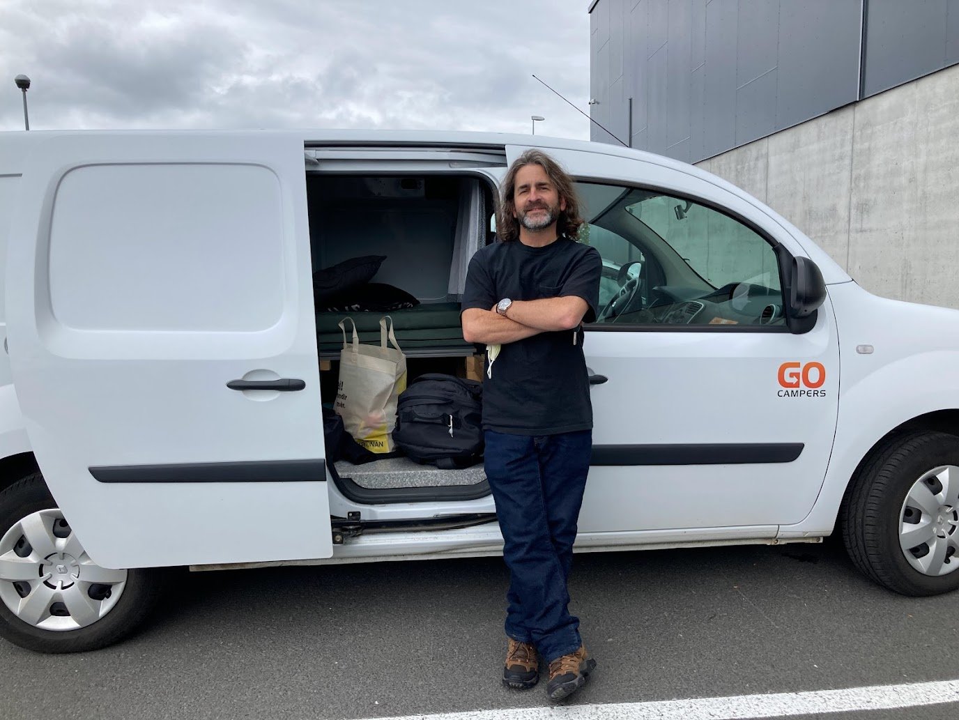 A man with long hair and a beard, wearing a black shirt and blue pants, stands with arms crossed in front of a GO Campers van—perfect for camping in Iceland—parked on a gray street near a concrete wall.
