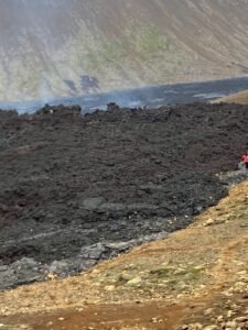 A rugged, dark lava field stretches across the landscape near the Ring Road, with faint steam rising and rocky earth bordering the scene. A person in a red jacket stands at the edge, capturing the wild spirit of camping in Iceland.