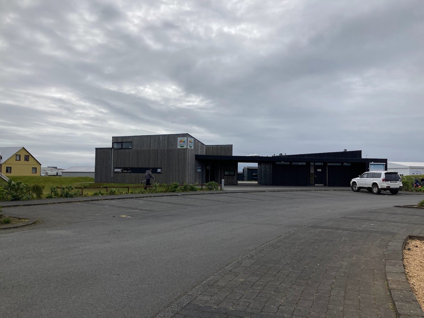 A modern, gray building with large windows sits beside a parking lot under a cloudy sky along the Ring Road. A person walks nearby, and a white SUV is parked to the right. In the background, there is a yellow house and open green fields.