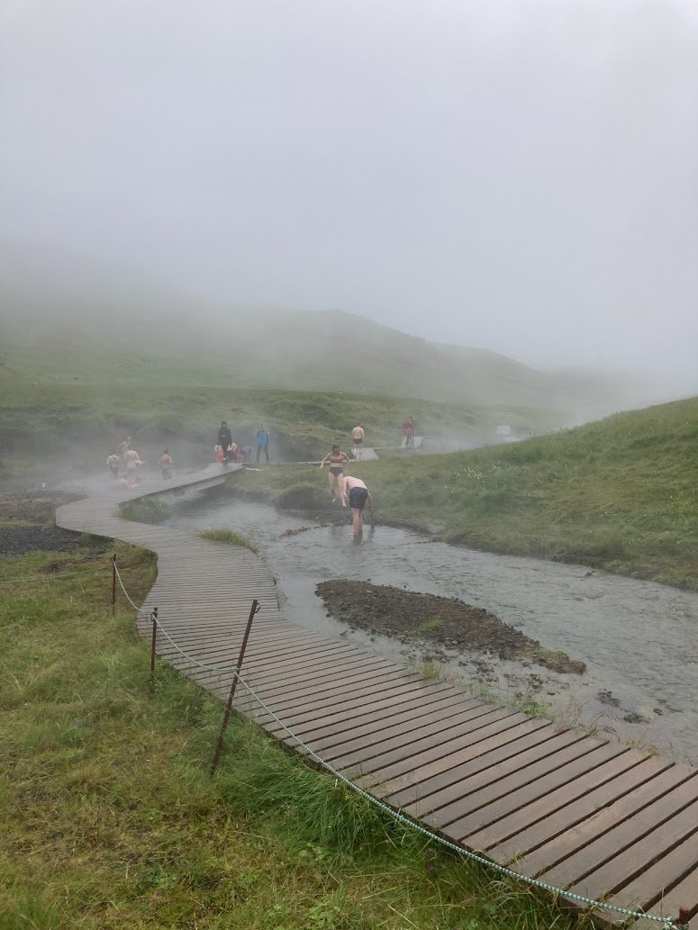 People bathe in a steamy hot spring alongside a winding wooden boardwalk in a misty, grassy landscape along Iceland’s famous Ring Road. The foggy scene, perfect for camping in Iceland in August, features hills rising in the background.