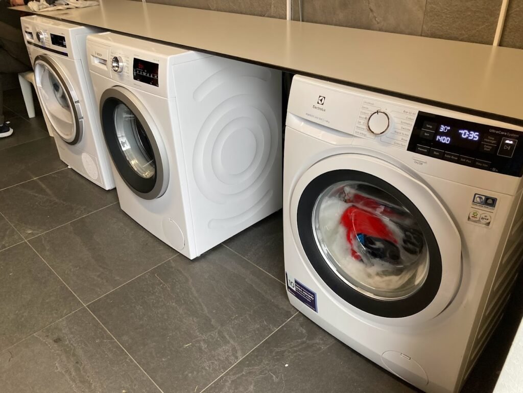 Three front-loading washing machines are lined up under a counter in a laundry room—perfect for freshening up after camping in Iceland. The machine on the right is running, with clothes inside the drum. The floor is tiled in dark gray.