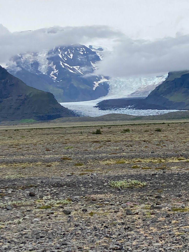 A rocky plain stretches toward distant snow-capped mountains, partially covered by clouds, with a glacier flowing down between the peaks—a stunning view often seen while camping in Iceland along the Ring Road.
