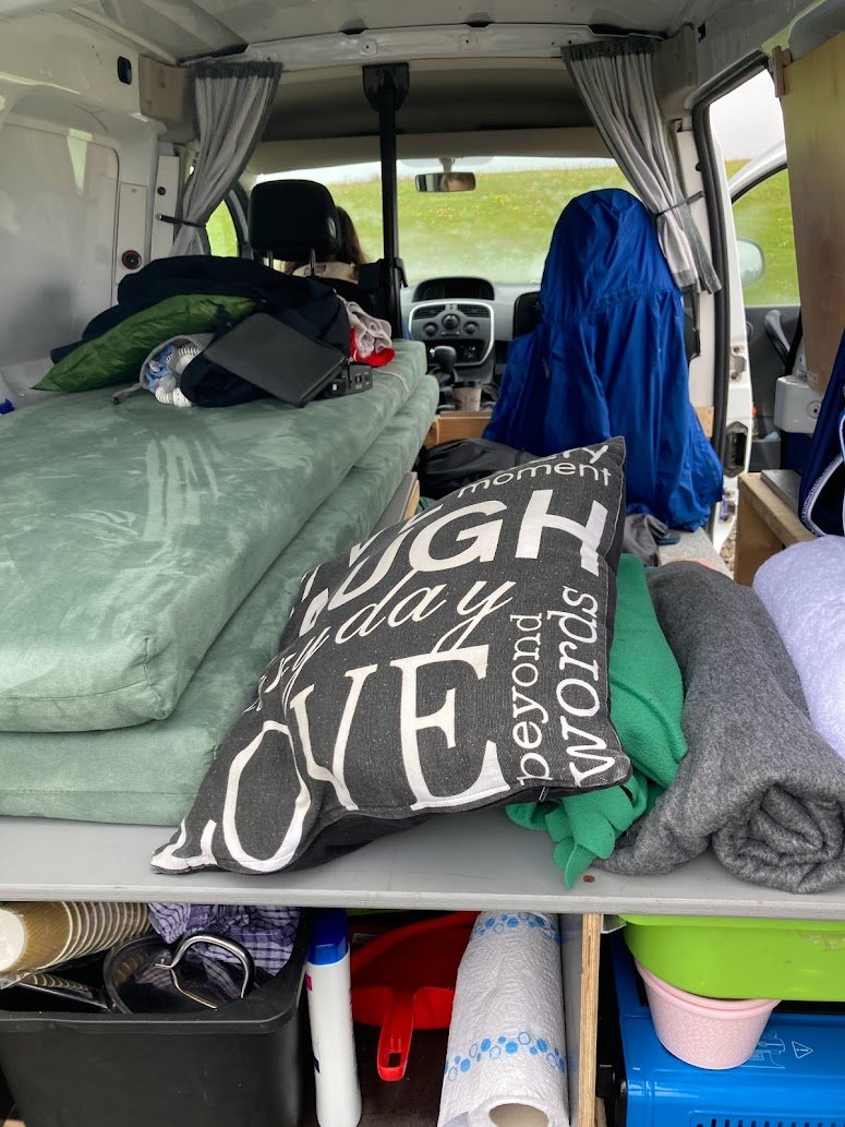 The interior of a camper van set up for camping in Iceland, with a made-up bed, pillows, blankets, a blue jacket, and supplies like towels and kitchen rolls neatly organized below the bed platform.