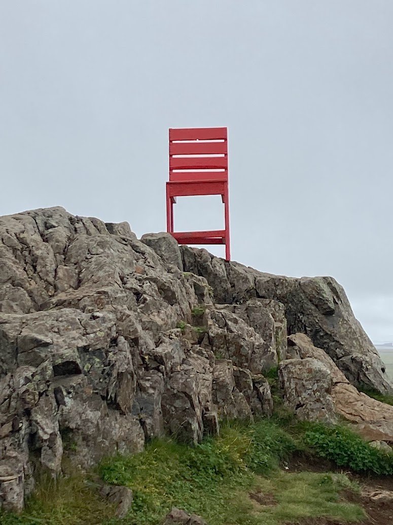 A large red wooden chair stands atop a rocky hill along Icelands Ring Road, appearing oversized against the rugged landscape—perfect scenery for camping in Iceland beneath cloudy skies and patches of green grass.