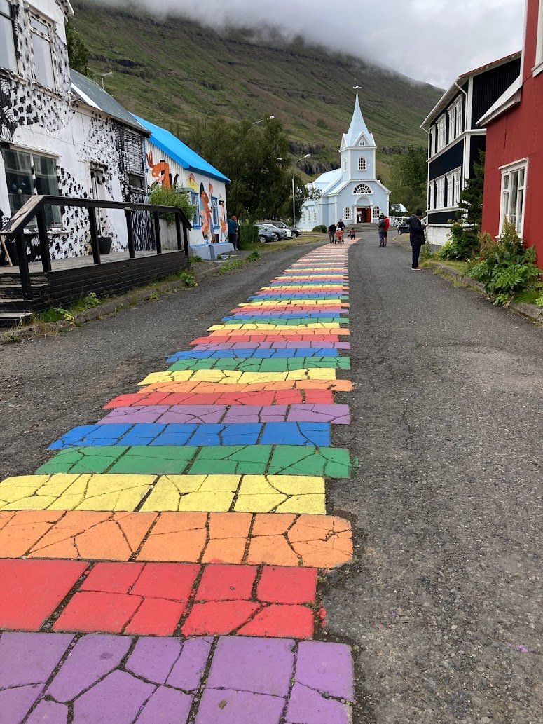A cobblestone street with rainbow-colored stones leads to a light blue church with a pointed steeple, surrounded by colorful buildings and mountains in the background.