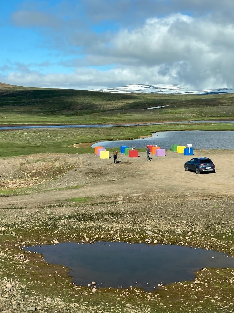 A grassy landscape along the Ring Road with a small pond, scattered rocks, a parked black car, and colorful cube-shaped structures near a river. Two people stand by the cubes under partly cloudy skies, with distant snow-capped hills—perfect for camping in Iceland in August.