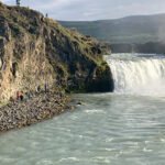 People stand on rocky terrain near the edge of a cascading waterfall, with mist rising and cliffs in the background under a cloudy sky—a breathtaking scene perfect for camping in Iceland.