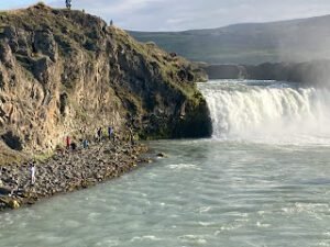 People stand on rocky terrain near the edge of a cascading waterfall, with mist rising and cliffs in the background under a cloudy sky—a breathtaking scene perfect for camping in Iceland.