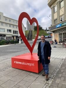 A person stands next to a large red heart sculpture with the hashtag #loveakureyri on a city street, with buildings, steps, and greenery visible in the background.