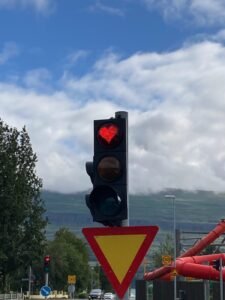 A traffic light shows a red heart-shaped signal above a yield sign, with a partly cloudy sky, trees, and a mountain in the background.