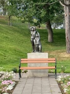 A bronze statue of two people with arms raised stands behind a park bench in a grassy area filled with trees and flower beds—an inviting spot to rest after camping in Iceland or exploring the scenic Ring Road.