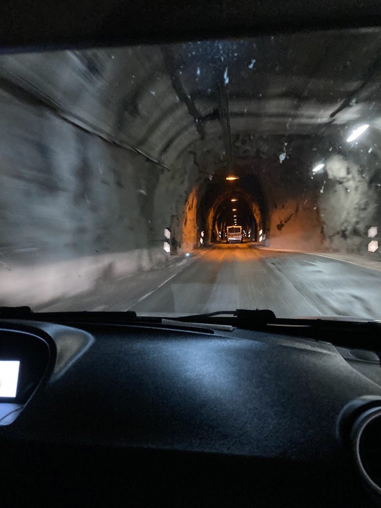 View from inside a car driving through a dimly lit tunnel with rough stone walls; another vehicle is visible ahead under yellowish lights.