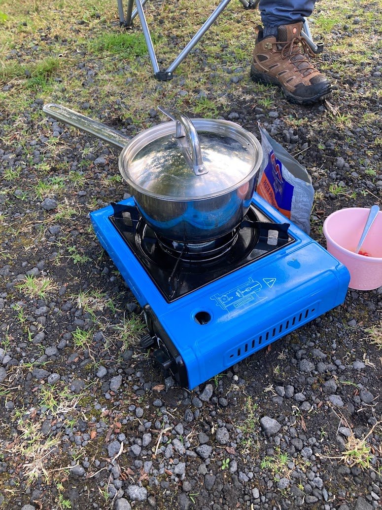 A blue portable camping stove with a pot and lid on top, set on gravel outdoors. A person’s hiking boot is visible, along with a pink bowl, spoon, and a food packet nearby. Grass is growing between the rocks.