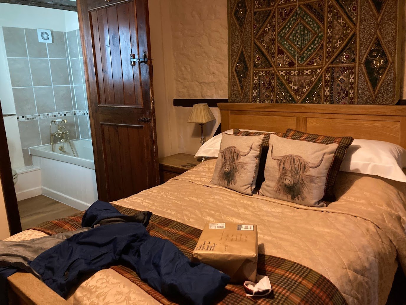 A cozy bedroom with a double bed, two pillows featuring highland cows, a brown blanket, a jacket and package on the bed, wooden furniture, and an open door leading to a tiled bathroom with a bathtub.