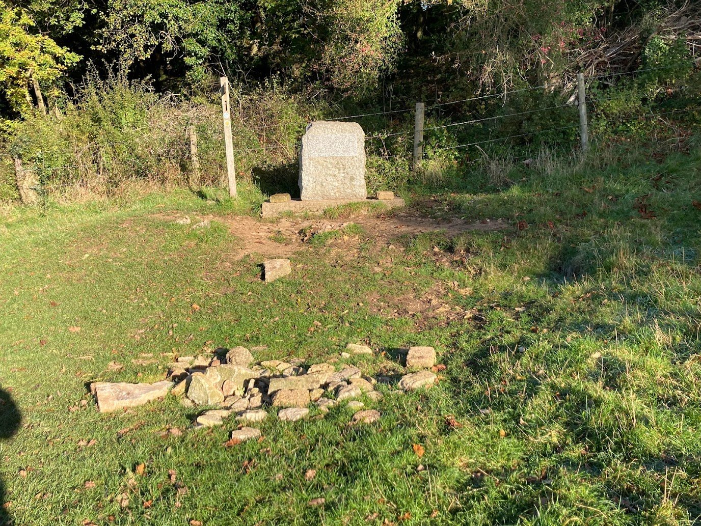 A stone monument stands on a grassy clearing along the Thames Path near a wire fence and trees, with scattered rocks on the ground in front of it, illuminated by sunlight.