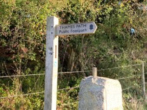 A wooden signpost points right, reading Thames Path Public Footpath with an acorn symbol. It stands next to a stone and wire fence, with greenery and sunlight in the background.