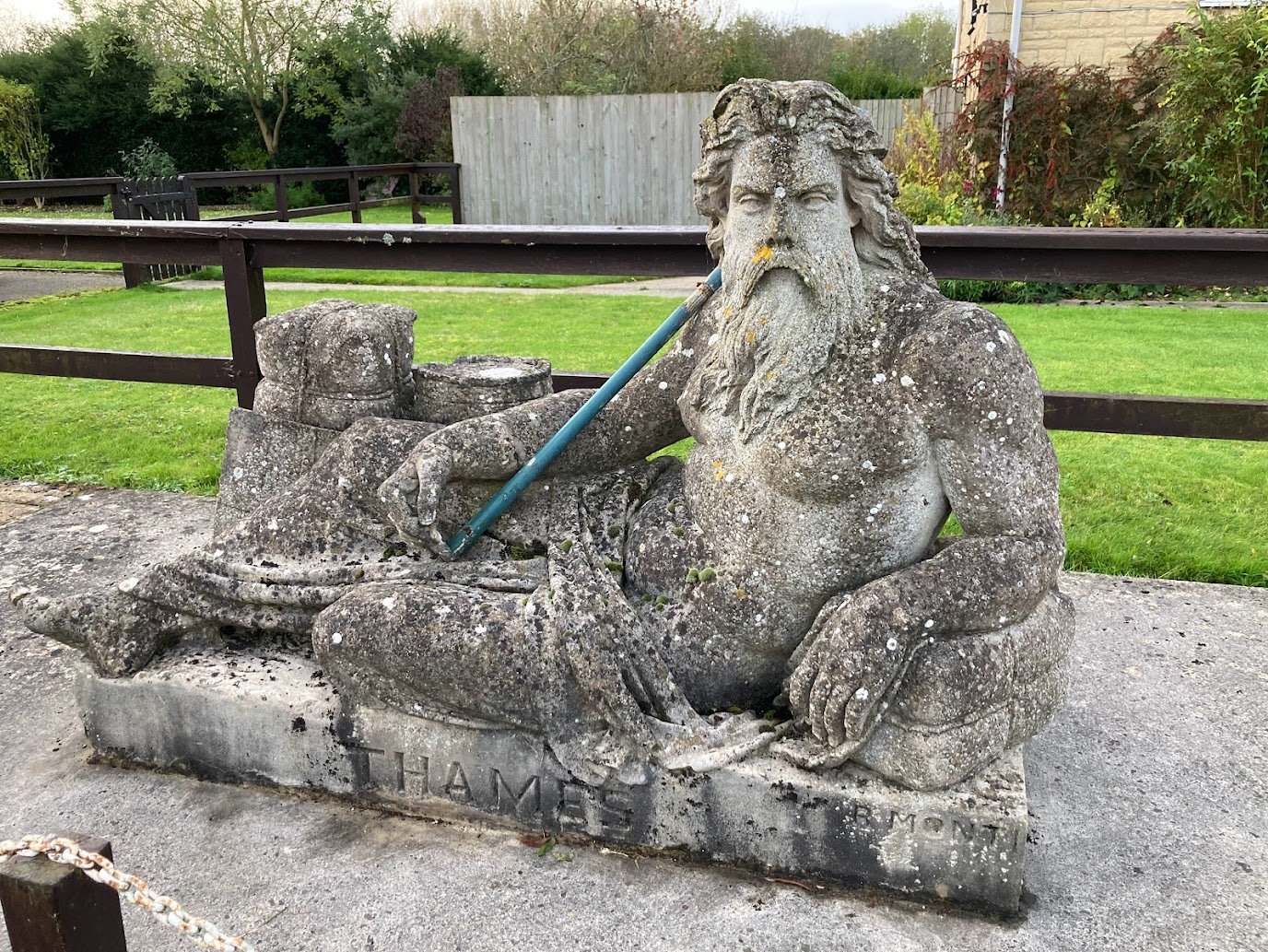 A weathered stone statue of a bearded, reclining man with flowing hair, draped in cloth, holding a staff. The base is inscribed with “THAMES.” The statue sits outdoors on a paved area with grass and a wooden fence behind.