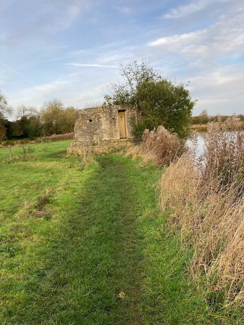 A narrow grassy path, part of the Thames Path, leads to an old stone building with a wooden door, surrounded by tall dry grass and bushes beside a calm river under a partly cloudy sky.
