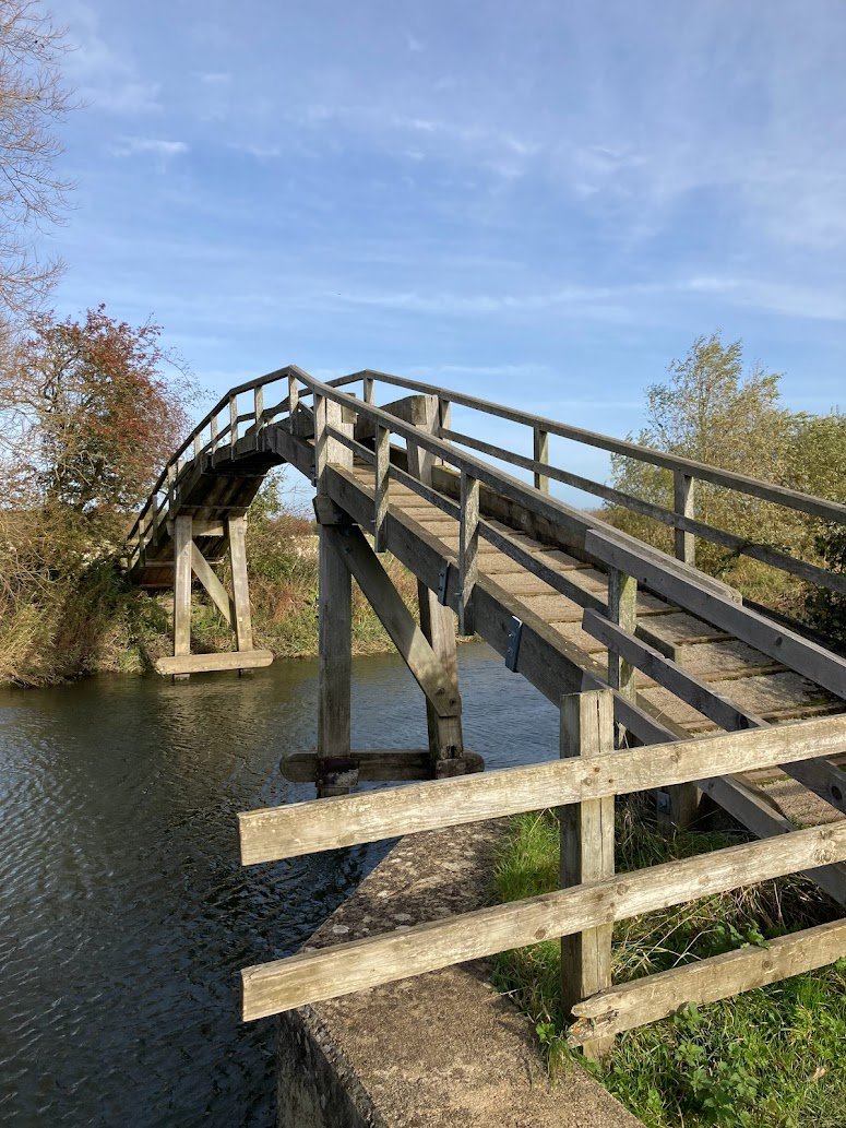 A wooden footbridge with railings arches steeply over a calm river, surrounded by trees and grassy banks under a clear blue sky.