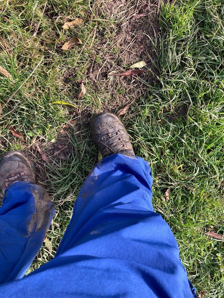 Person wearing blue pants and muddy black shoes standing on green grass with patches of dirt and fallen leaves visible. Photo is taken from the persons perspective, looking down at their legs and feet.