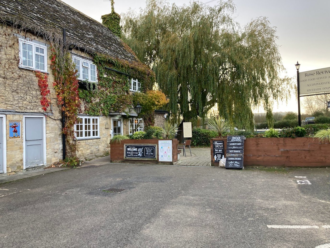A rustic stone building with ivy and red vines on the walls, featuring white-framed windows, stands near outdoor signs and menu boards along the Thames Path. A large willow tree and a sign for Rose Revived pub are visible in the background.