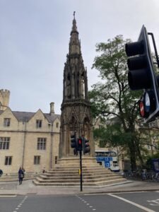 A tall, ornate stone monument with gothic details stands on a stepped base at a street corner along the Thames Path, surrounded by historic buildings, trees, traffic lights, and pedestrians.